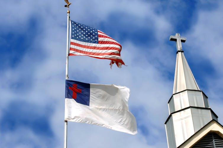 A white flag with a red cross in the corner flies below an American flag, next to a church steeple.