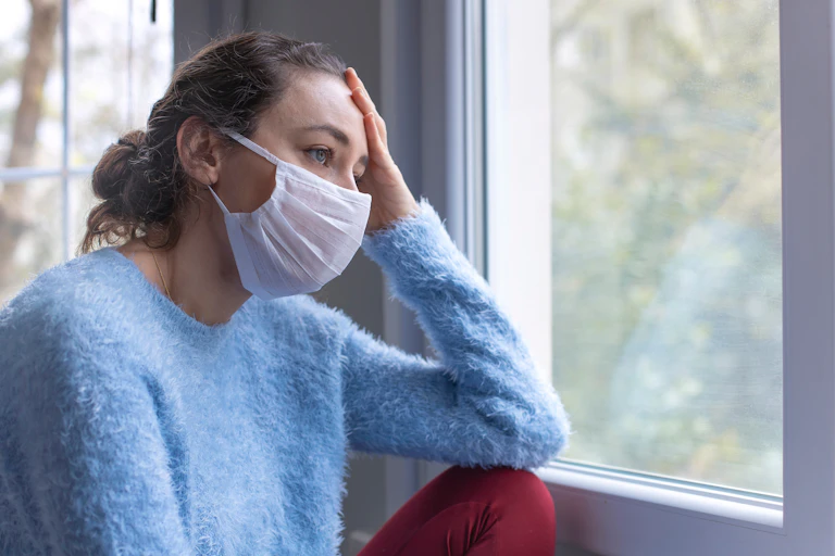 A young woman in a blue sweater sitting a window wearing a face mask looking tired