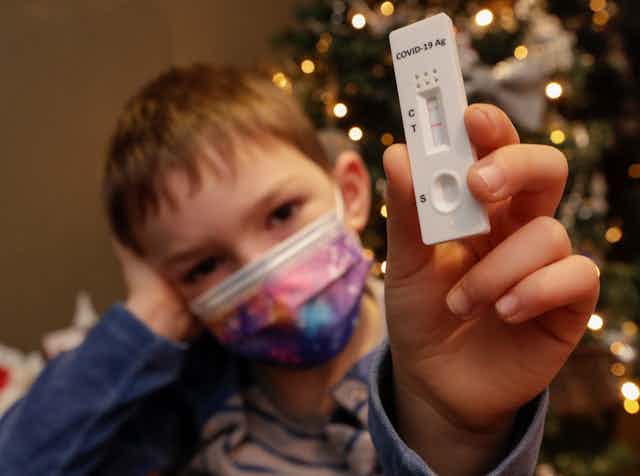 A child wearing a mask holding up a positive rapid antigen test