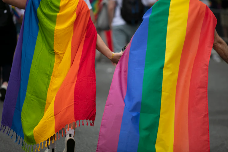 Two men wearing gay pride flags as capes, holding hands and walking away from the camera
