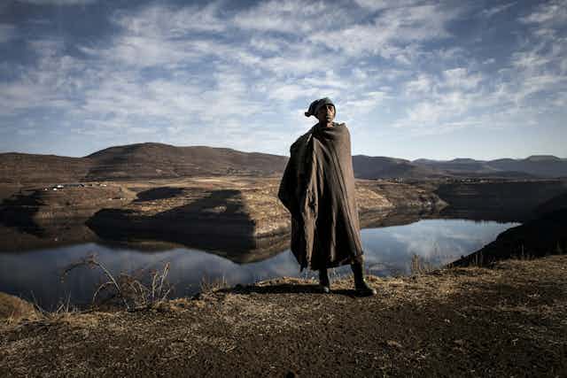 A man stands in front of mountains and a dam, dressed in a blanket, boots and a cap.