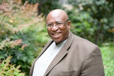 A bald man in a suit jacket stands in a garden smiling at the camera.