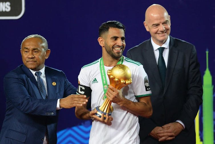 Un joueur de football tient une coupe dorée entre ces mains, entouré par deux hommes en costume. Tous sourient.
