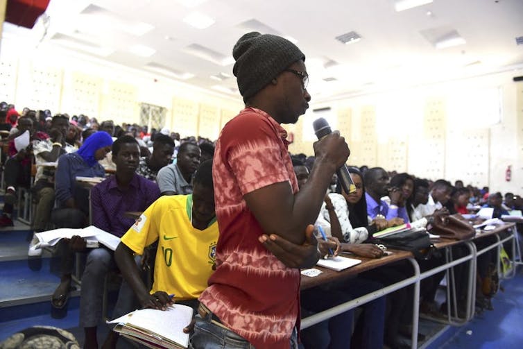 Un étudiant avec un micro en main parle devant un amphithéâtre bondé.