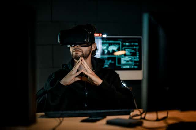 A man wearing virtual reality goggles in a dark room with a computer screen behind him