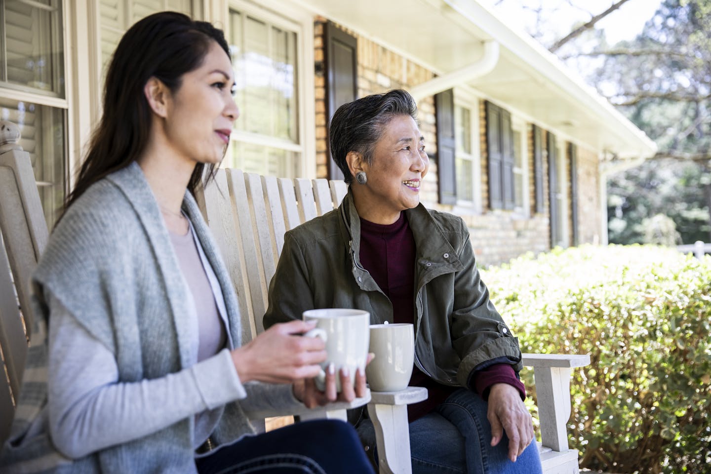 A woman and her older mother sit on a porch, drinking coffee.