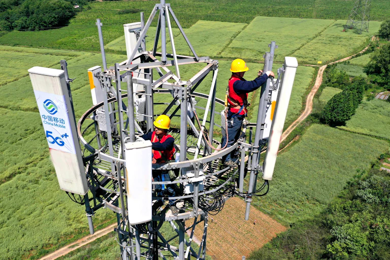 Two people wearing safety helmets, vests and harnesses work at the top of a tall telecommunications tower