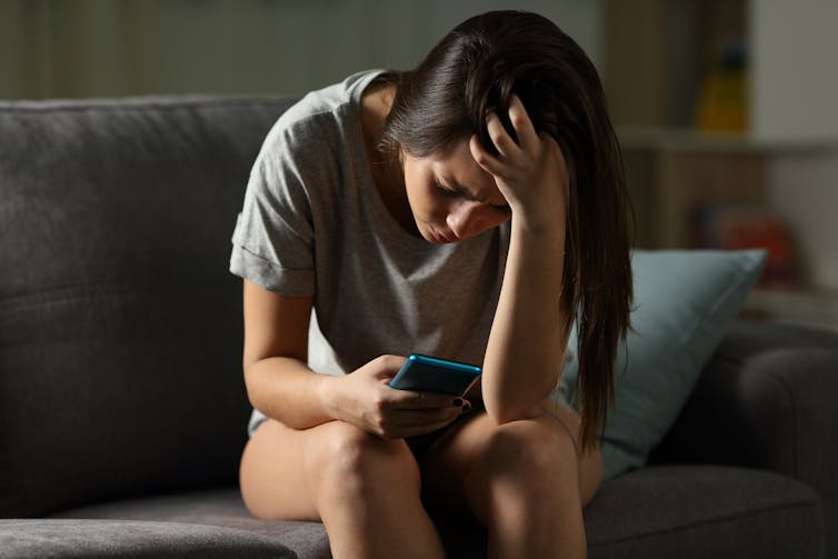 A woman sitting on a sofa, holding a cell phone in one hand and holding her head in distress with the other.