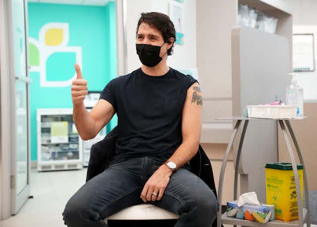 A man wearing a black t-shirt and black mask gives the thumbs up after receiving a vaccine in his left arm