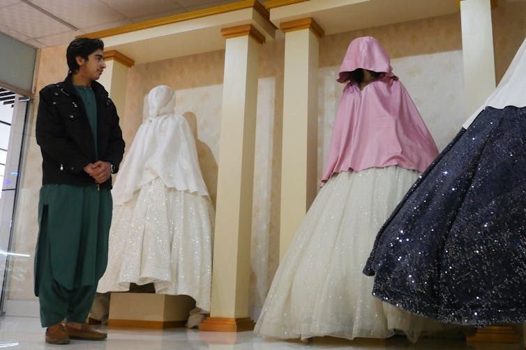 A shopkeeper stands in front of three mannequins that are covered from head to toe with draping clothing.