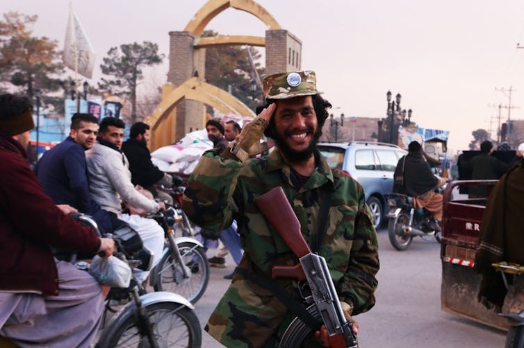 A Taliban soldier salutes, smiling, in front of a busy street, with men riding bikes and cars passing by.