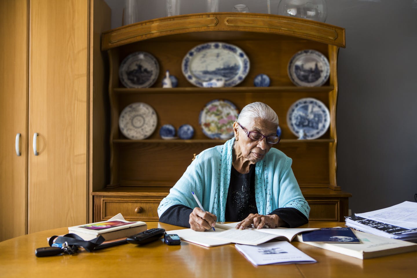 An older Black woman writes as she sits at a dining room table in front of a china cabinet.