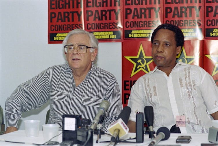 Two men field questions at a press conference while seated with their backs to Communist Party posters