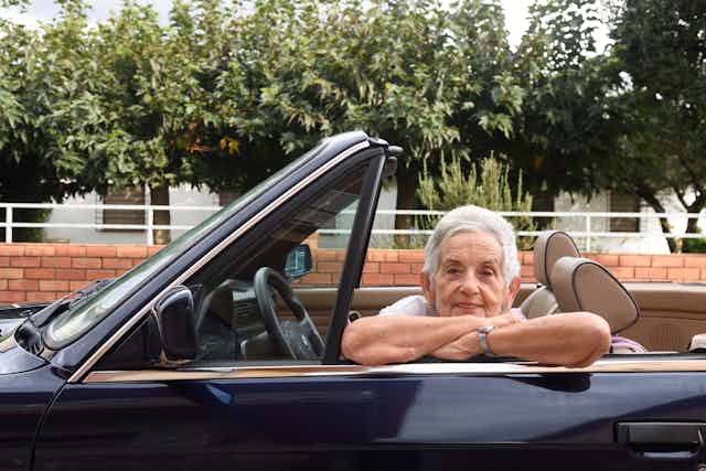 An elderly woman in the driver's seat of a convertible leans her arms on the door frame. 