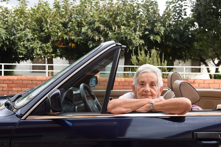An elderly woman in the driver's seat of a convertible leans her arms on the door frame.