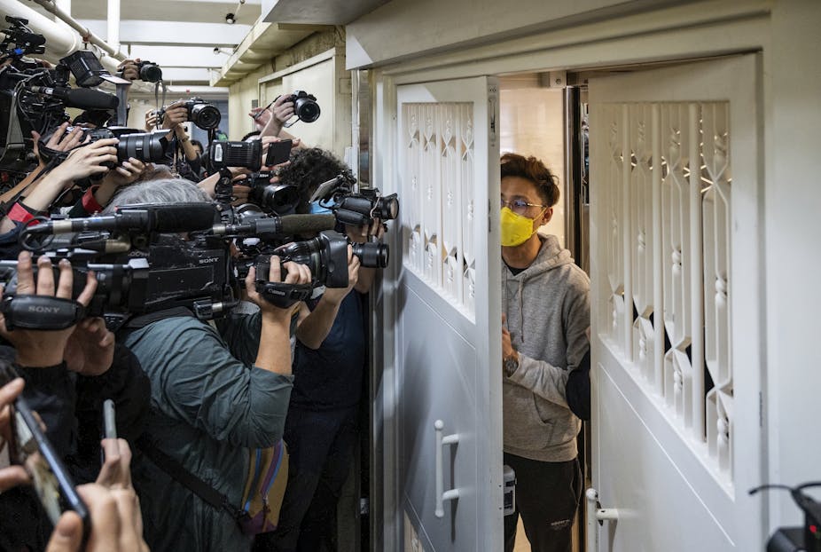 A young man in a yellow face mask and glasses approaches a doorway, outside which a crowd of media cameras are waiting