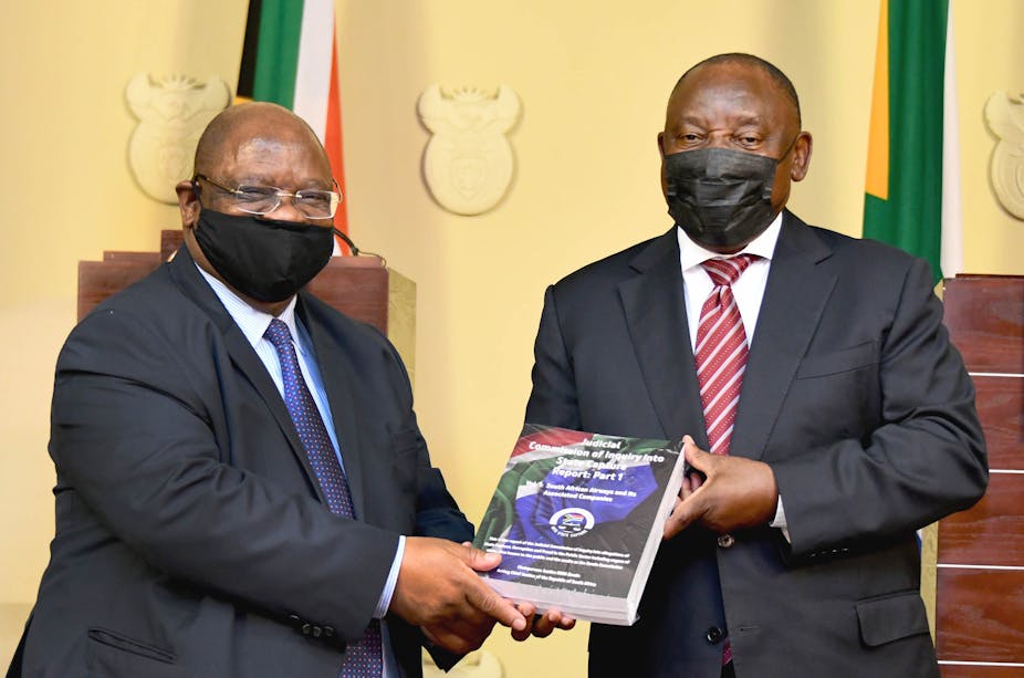 A bald-headed man wearing a suit hands over a thick report to another, also wearing a suit, in front of two South African flags. Both men are wearing Covid-19 masks.