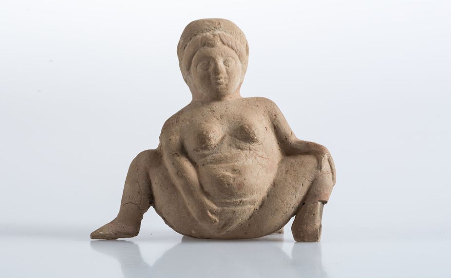 Estatuilla de mujer desnuda abriendo las piernas y mostrando su vulva con la mano derecha.