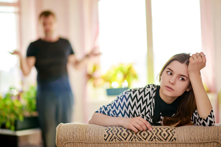 A girl sits with her hand on her head as she ignores a lecturing parent blurred behind her.