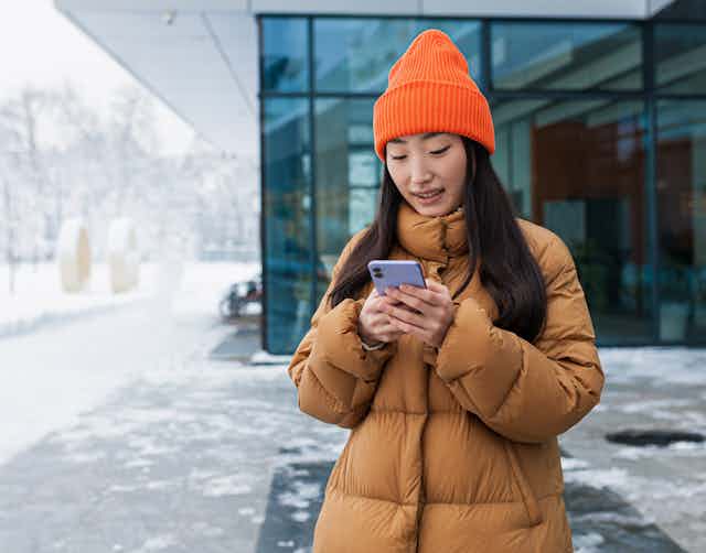 A woman in a winter coat and orange hat standing outside reading a text message.