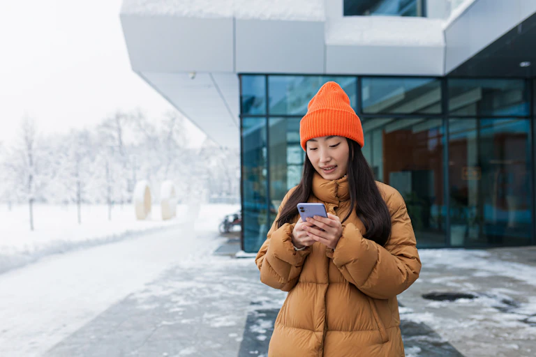 A woman in a winter coat and orange hat standing outside reading a text message.