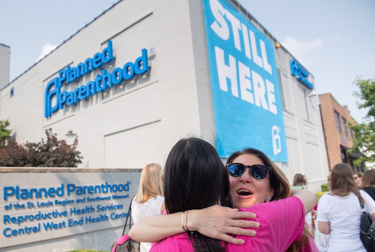 Two women hug outside of a Planned Parenthood clinic, which has a sign that says 'still here' on the side