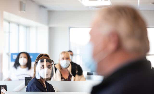 Doug Ford in the foreground as a health-care worker in a viser and other people in masks look at him.