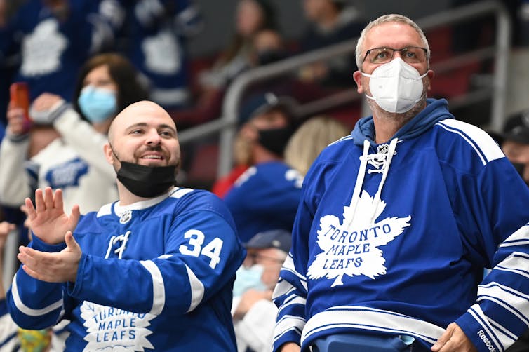Two men in Leafs jerseys, one with his mask under his chin and the other with a mask over his mouth and nose.