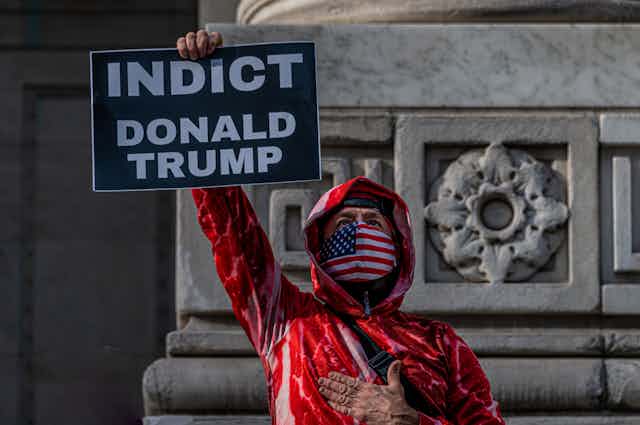A man dressed in a red jacket, with a US flag mask, holding a sign that says "INDICT DONALD TRUMP"