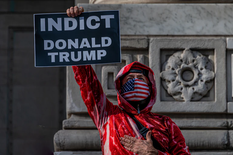 A man dressed in a red jacket, with a US flag mask, holding a sign that says "INDICT DONALD TRUMP"