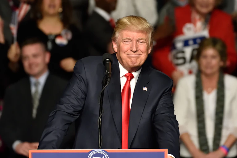 Donald Trump stands behind a podium wearing a navy suit with a smug grin on his face.