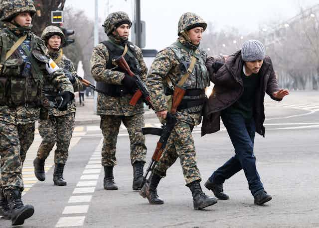 Military forces dressed in fatigues and holding guns hold a man's arm behind is back.