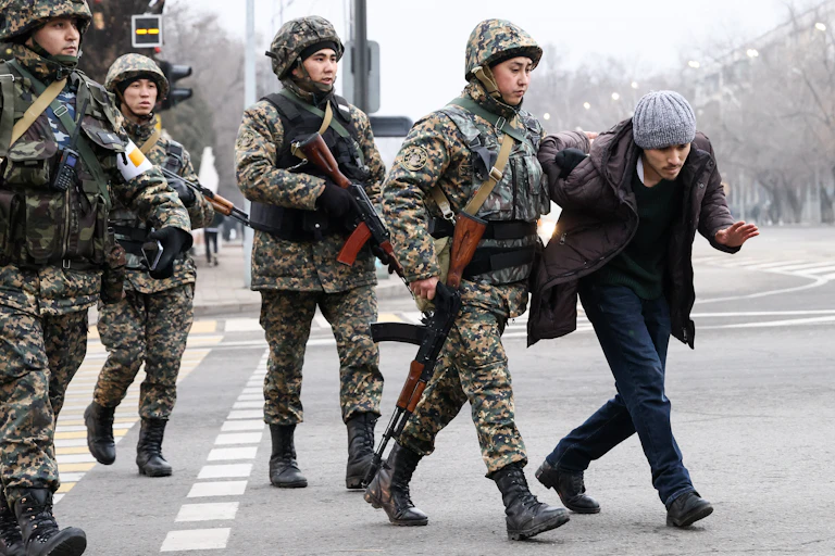 Military forces dressed in fatigues and holding guns hold a man's arm behind is back.