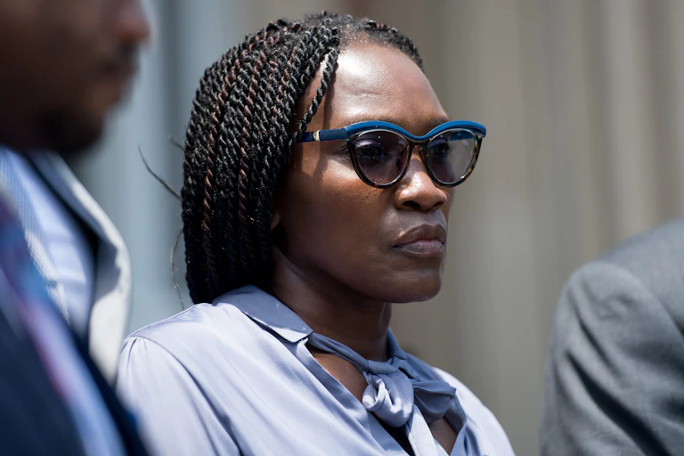 A Black woman in sunglasses is flanked by two men in suits.