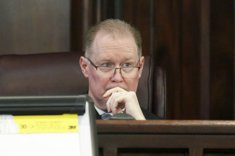 A pensive judge looks out into a courtroom with his hand pressed to his mouth.