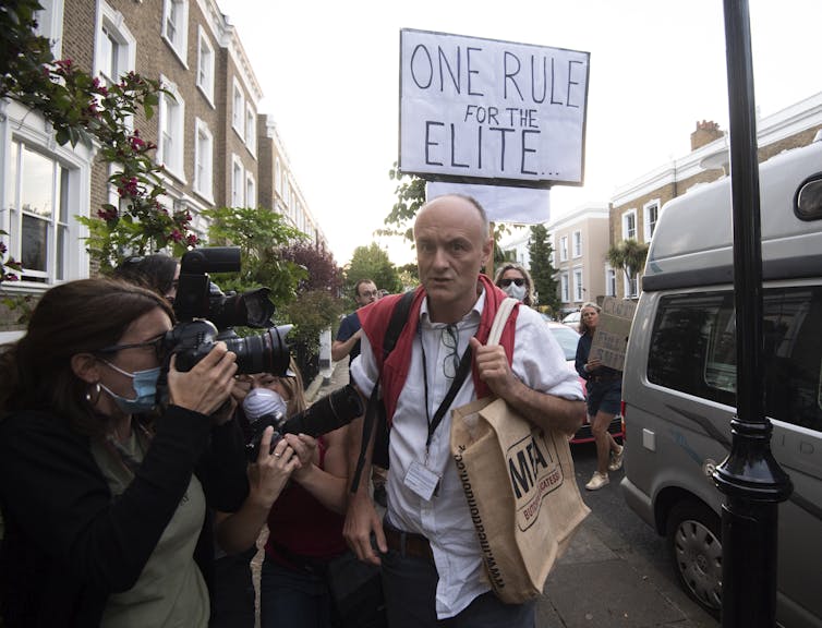 Dominic Cummings swamped by media and protestors, one holding a sign behind him that reads 'One rule for the elite'/