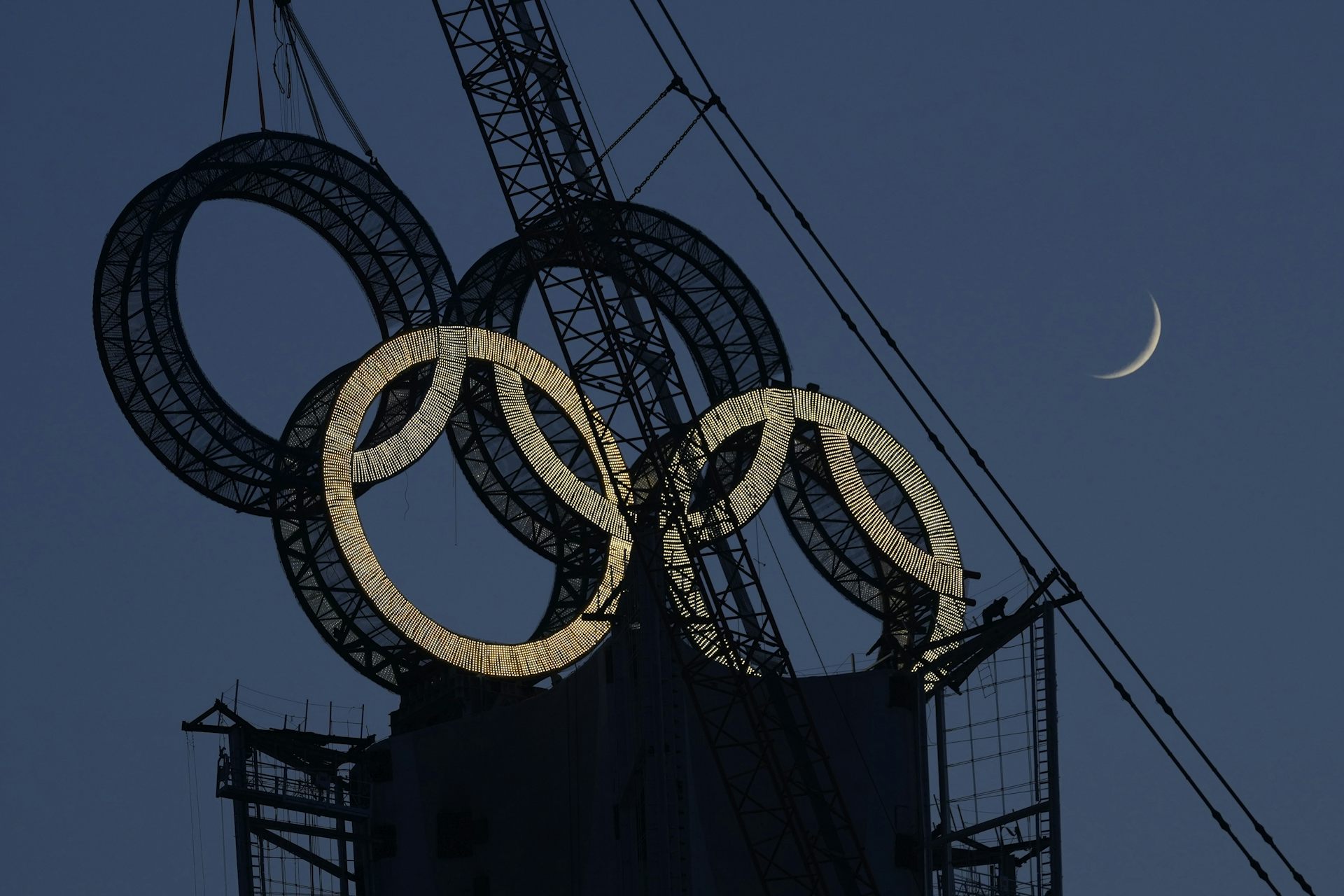 A worker works to assemble the Olympic Rings onto a tower on the outskirts of Beijing. In the photo you can see a crane, four rings and a cresent moon in the sky.