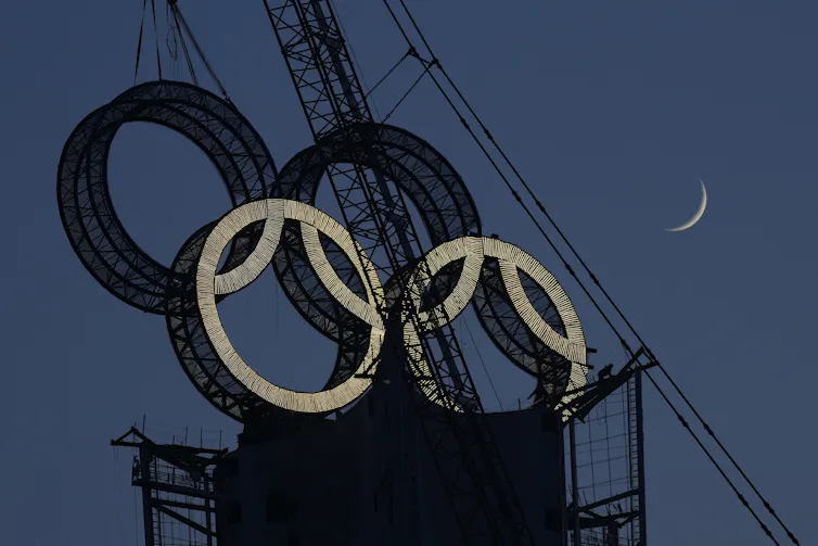 A worker works to assemble the Olympic Rings onto a tower on the outskirts of Beijing. In the photo you can see a crane, four rings and a cresent moon in the sky.