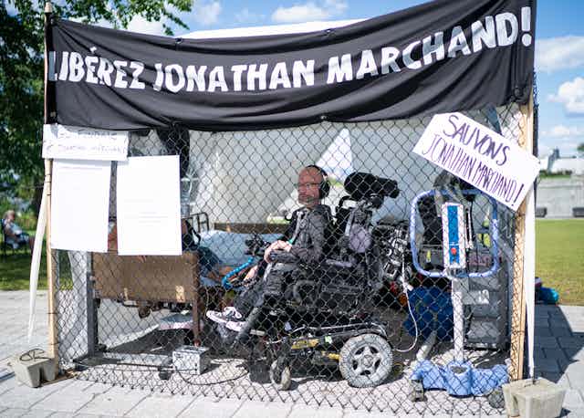 A man sits in a cage near the Quebec legislature he's in a wheelchair. A sign on top of the cage reads 'libérez jonathan marchand'