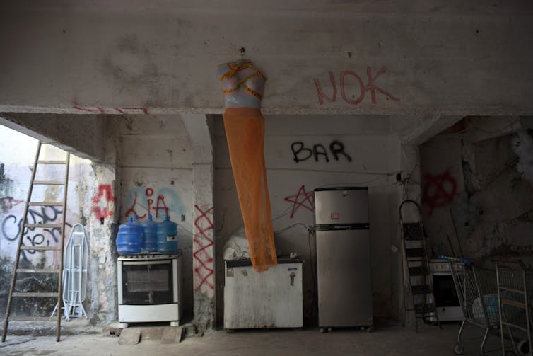 A half torso mannequin decorated with an orange veil hangs from a beam backdropped by donated items, a stove and fridge. The room is covered in grafitti.