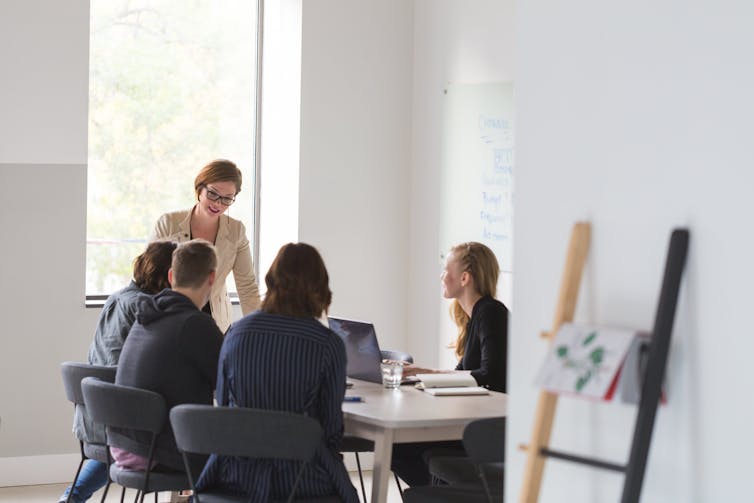 A woman stands at the head of a table in a meeting room with white walls and a window behind her talking to her team.