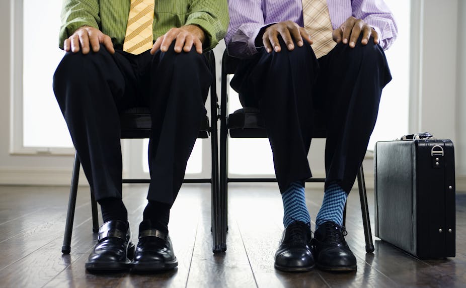 Two men sit side by side, visible from the waist down, one Black, one white, one with black socks and the other with colorful socks.