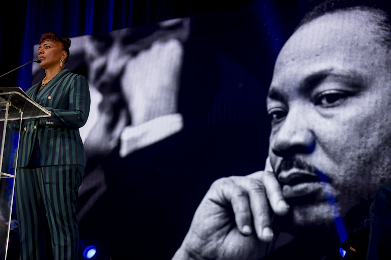 A woman wearing suit pants speaks at a podium next to the image showing the face of a man in a contemplative mood, with his right hand on his face.