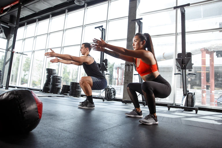A young man and woman in gym clothes perform a bodyweight squat in a gym.