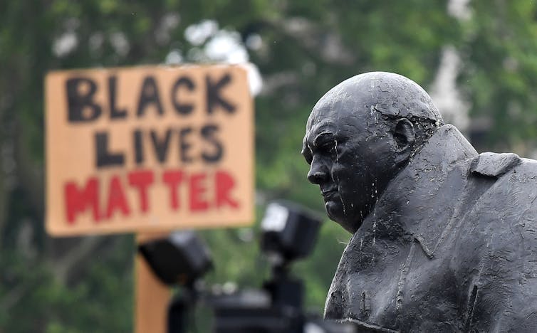 Χάλκινο άγαλμα του Ουίνστον Τσόρτσιλ σε πρώτο πλάνο, μια πινακίδα Black Lives Matter στο βάθος.