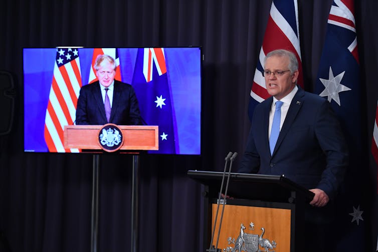 Australian PM Scott Morrison speaks at a podium while Boris Johnson does the same on a screen beside Morrison.