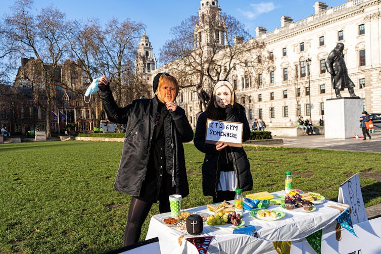 Two protestors hold up wine and nibbles in Whitehall whilst wearing masks depicting Boris and Carrie Johnson. One holds a sign reading 'it's 6pm somewhere'.
