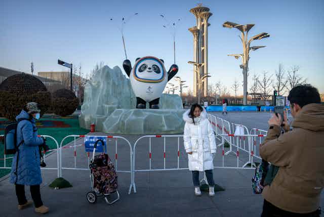 A woman poses in front of a statue of an Olympic mascott. She's wearing a big white jacket.