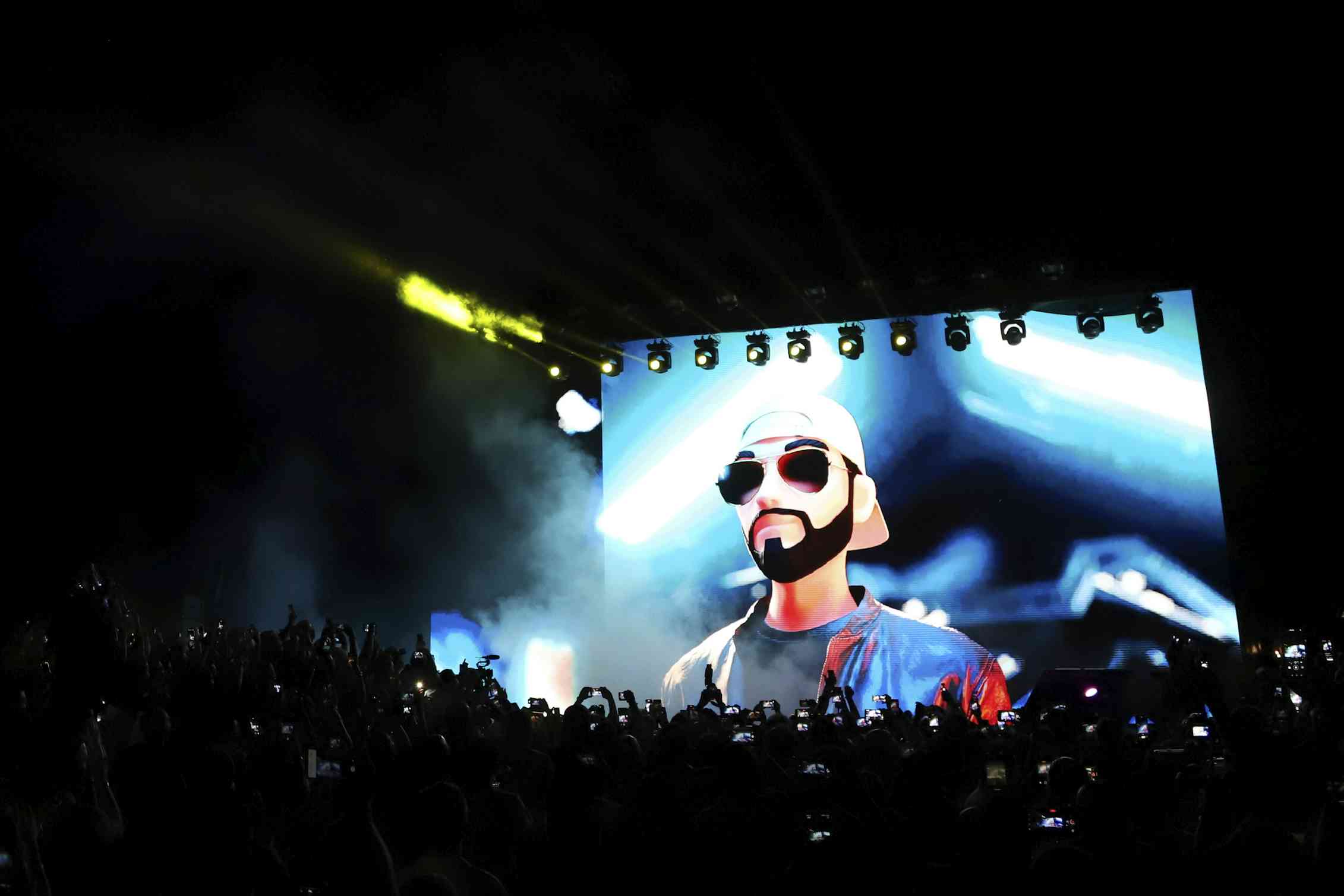 a cartoon image of a young bearded man wearing sunglasses and a backwards ball cap is projected on a wall in a darkened auditorium