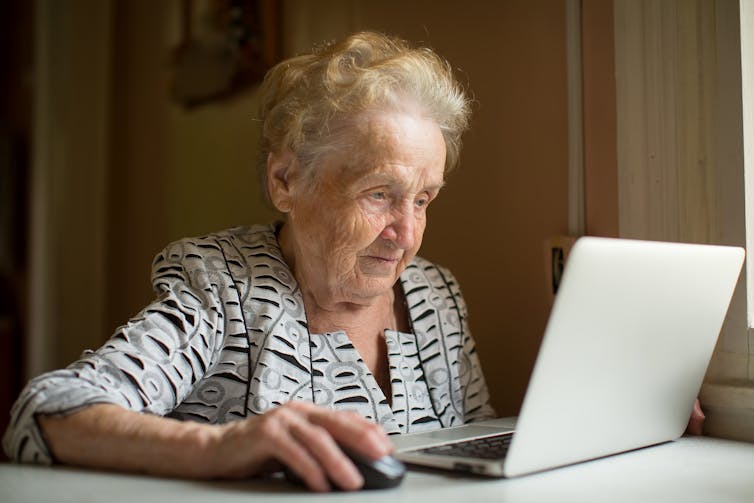 An older woman sits on her laptop. She is wearing a striped blouse, has short hair and us using a computer mouse.